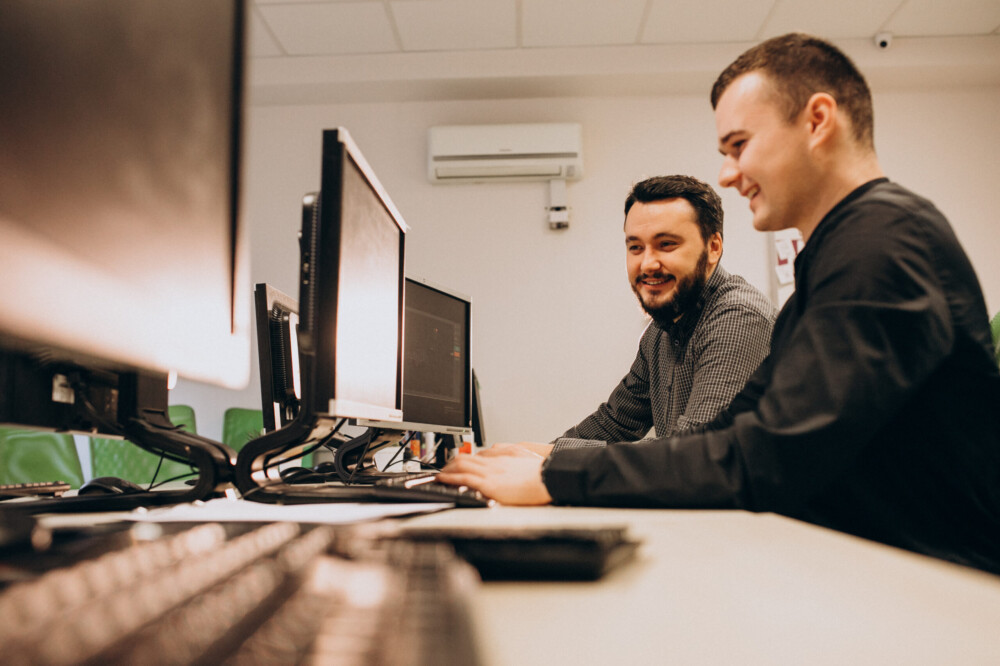 Zwei Männer sitzen an einem Schreibtisch mit PCs und lächeln